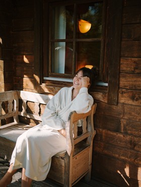 Woman in a bathrobe sitting on a wooden bench in front of a wooden hut