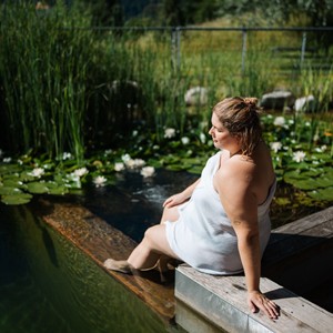 A woman in a towel sits on the jetty of a pond and dips her feet in the water.