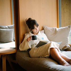 Woman in a bathrobe on a lounger reading a book with a cup in her hand