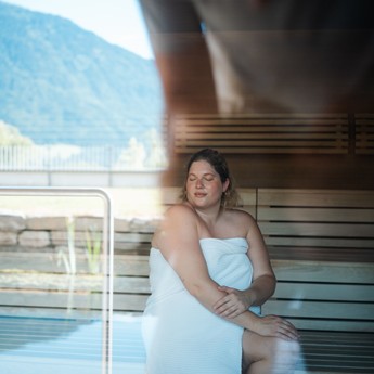 Eine Frau in der Sauna mit Panoramafenster, in der Spiegelung ist der Außenbereich zu sehen.