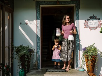 Frau hält zwei Kannen Milch, Kind neben ihr trinkt aus einem Glas Milch