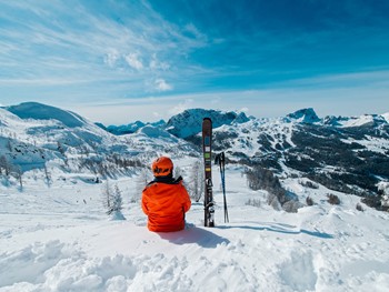 Person with skis next to them sits on a slope and looks into the mountain landscape