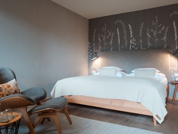 Gemütlich schlafen im Doppelzimmer Natur Doppelbett im Hotelzimmer mit grauem Kopfteil und einer dezenten Pflanzen-Tapete an der Wand dahinter.