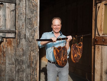 Lernen Sie richtiges Einsuren und Räuchern mit dem Gailtaler Speckmeister Mann hält eine Stange mit zwei großen, gereiften Stücken Fleisch