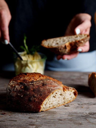 Angeschnittener Laib Brot, im Hintergrund wird eine Scheibe davon bestrichten